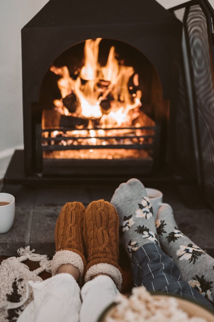 Paits of feet wearing socks and slippers are seen on a footstool in front of an open fire. Photo by Taryn Elliott on Pexels.