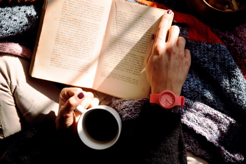 A person with painted nails and a pink watch holds a cup in one hand and a book on their lap in the other. Photo by Vincenzo Malagoli on Pexels