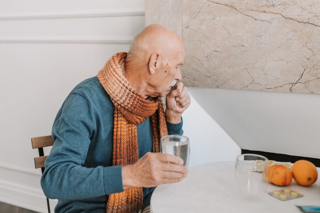 An older bald man with a moustache coughs. He is wearing a scarf and holding a glass of water. Oranges and lozenges are seen on the table in front of him. Photo by Vlada Karpovich on Pexels